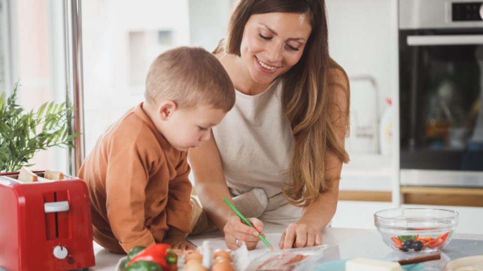 A mother and her young son writing a list at a kitchen counter with eggs, peppers and bacon beside them
