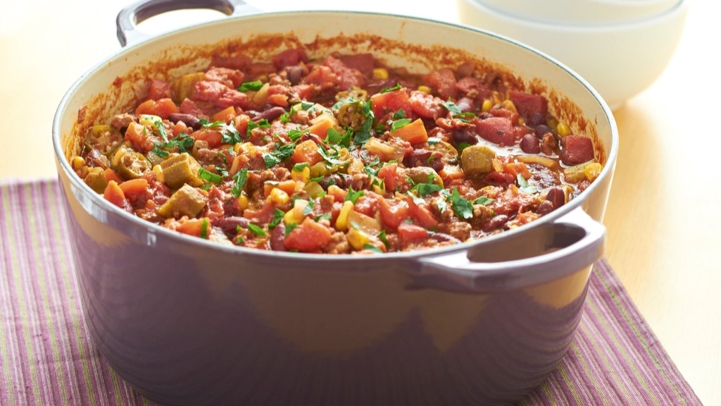 A large pot of chili with beans, corn and tomatoes, with a stack of bowls behind it
