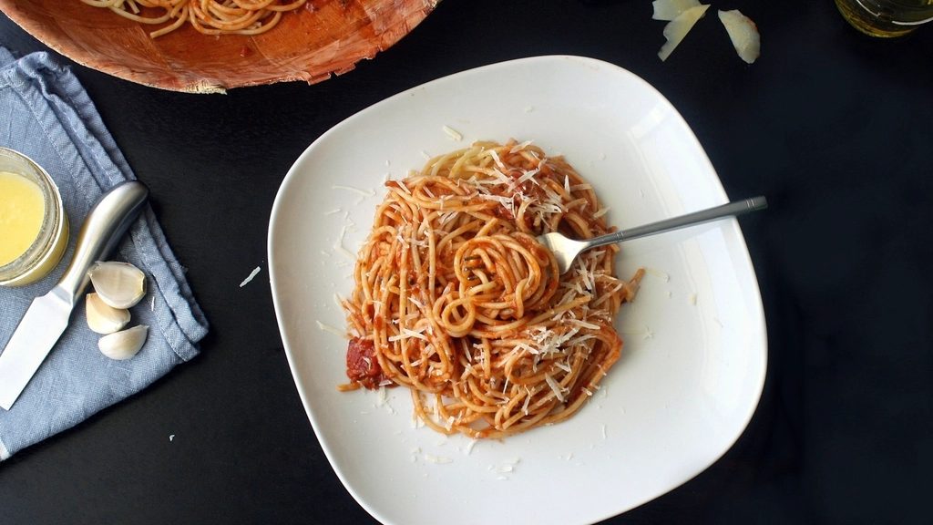 A plate of spaghetti in tomato sauce with parmesan, beside a large serving bowl of pasta, garlic and a grater
