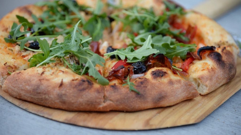A homemade pizza with arugula, olives and red peppers on a wooden pizza peel